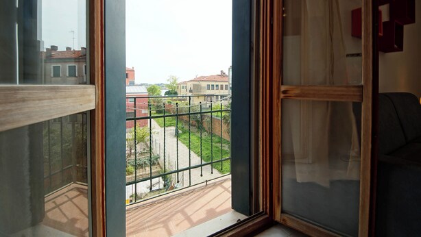 Ein Zimmer mit großem Fenster, das auf einen Innenhof mit Metallzaun, ein Kachelboden und eine Ansicht auf Gebäude in der Ferne blickt.