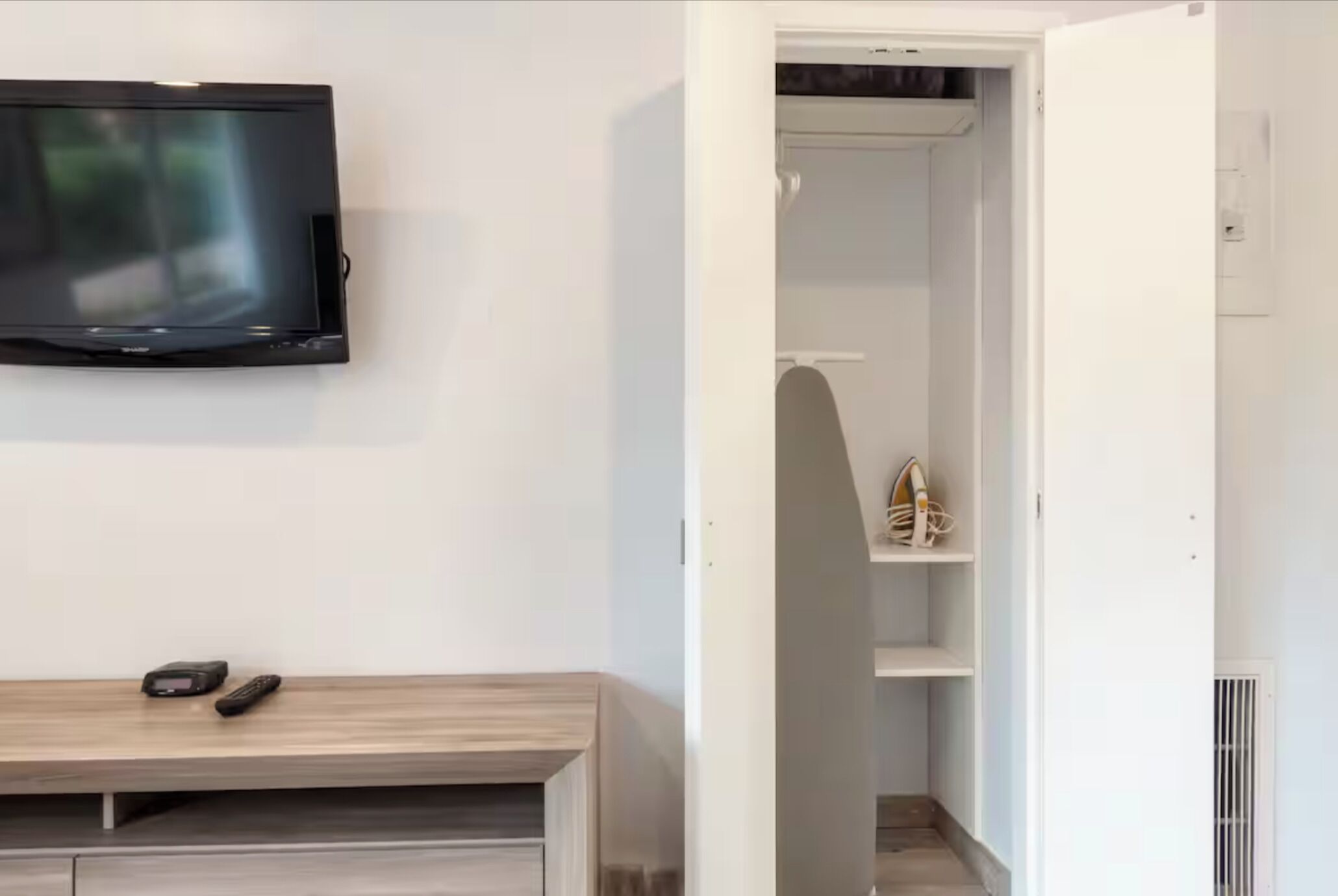 A room with a TV mounted on the wall, a wooden bench with shoes, and a closet with shelves.