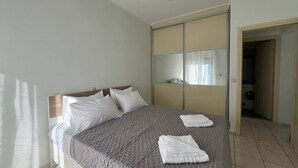 Una cama bien hecha con colcha gris, almohadas blancas y dos toallas dobladas. Hay un espejo grande en la pared y una puerta que da a un área de lavandería.