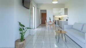 Una sala de estar moderna con un sofá blanco, una mesa de centro y un televisor montado en la pared. Hay una zona de comedor con una mesa y sillas, y una cocina al fondo.