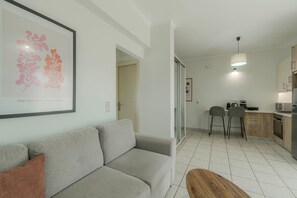 A modern living room with a grey sofa, a wooden coffee table, and a kitchen area with a dining table, chairs, and appliances.