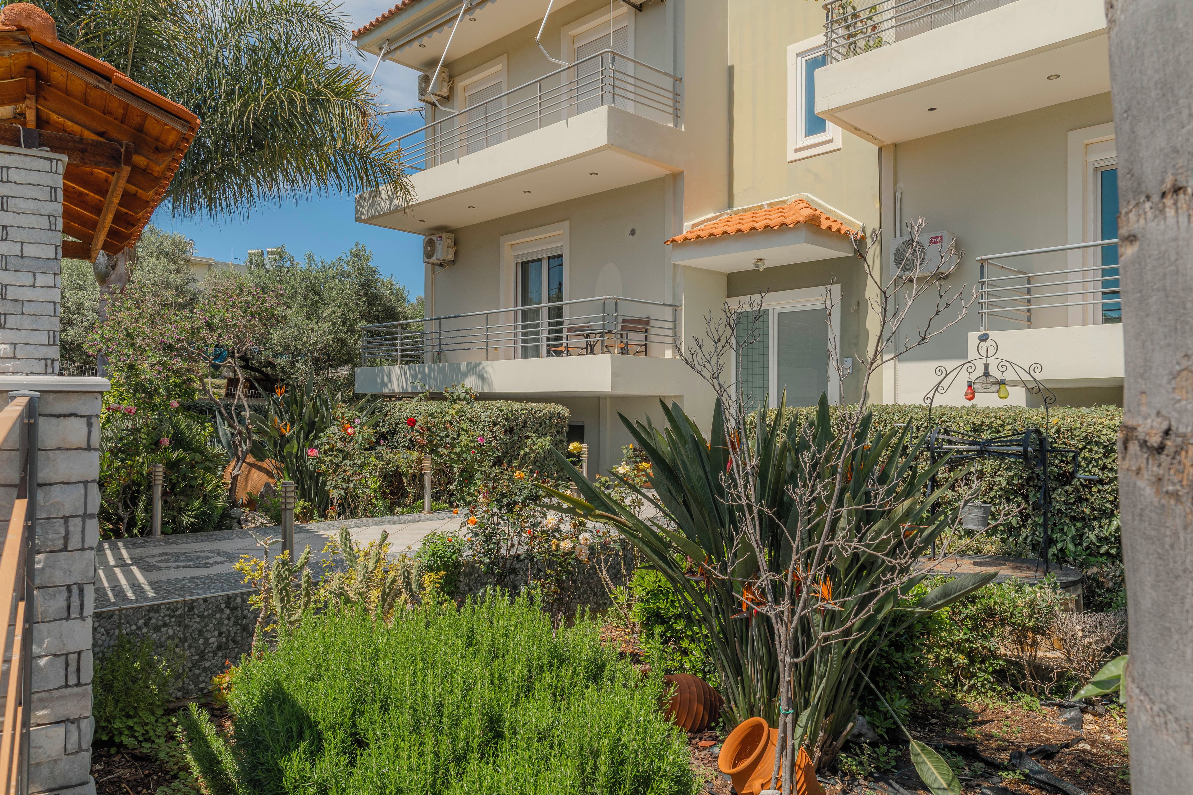 A multi-story residential building with balconies, a garden with various plants, and a clear sky.