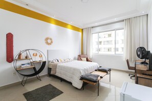 A modern bedroom with a bed, a desk, and a chair. There is a window with curtains, a radiator, and a circular shelf with books. - Coliving São João (São Paulo)