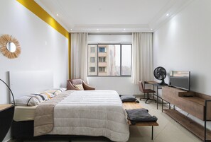 A modern hotel room with a large bed, a desk with a computer, a TV, and a view of the city through the window. - Coliving São João (São Paulo)