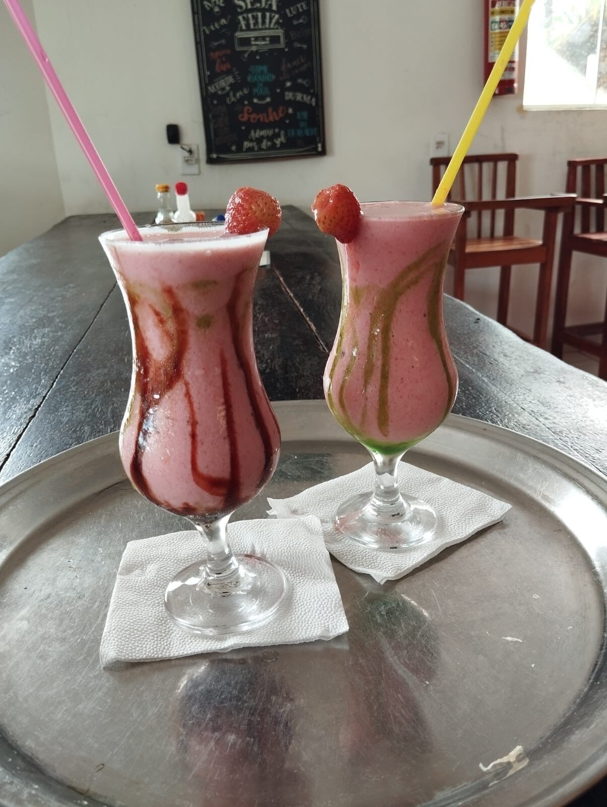 Two glasses of pink beverage on a metal tray with straws, placed on a table with a chalkboard in the background.