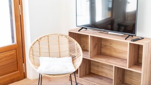 A room with a wooden TV stand, a flat-screen TV, a wicker chair, and a white cushion. - Casa Tharros Apartment Garden (Nurachi)