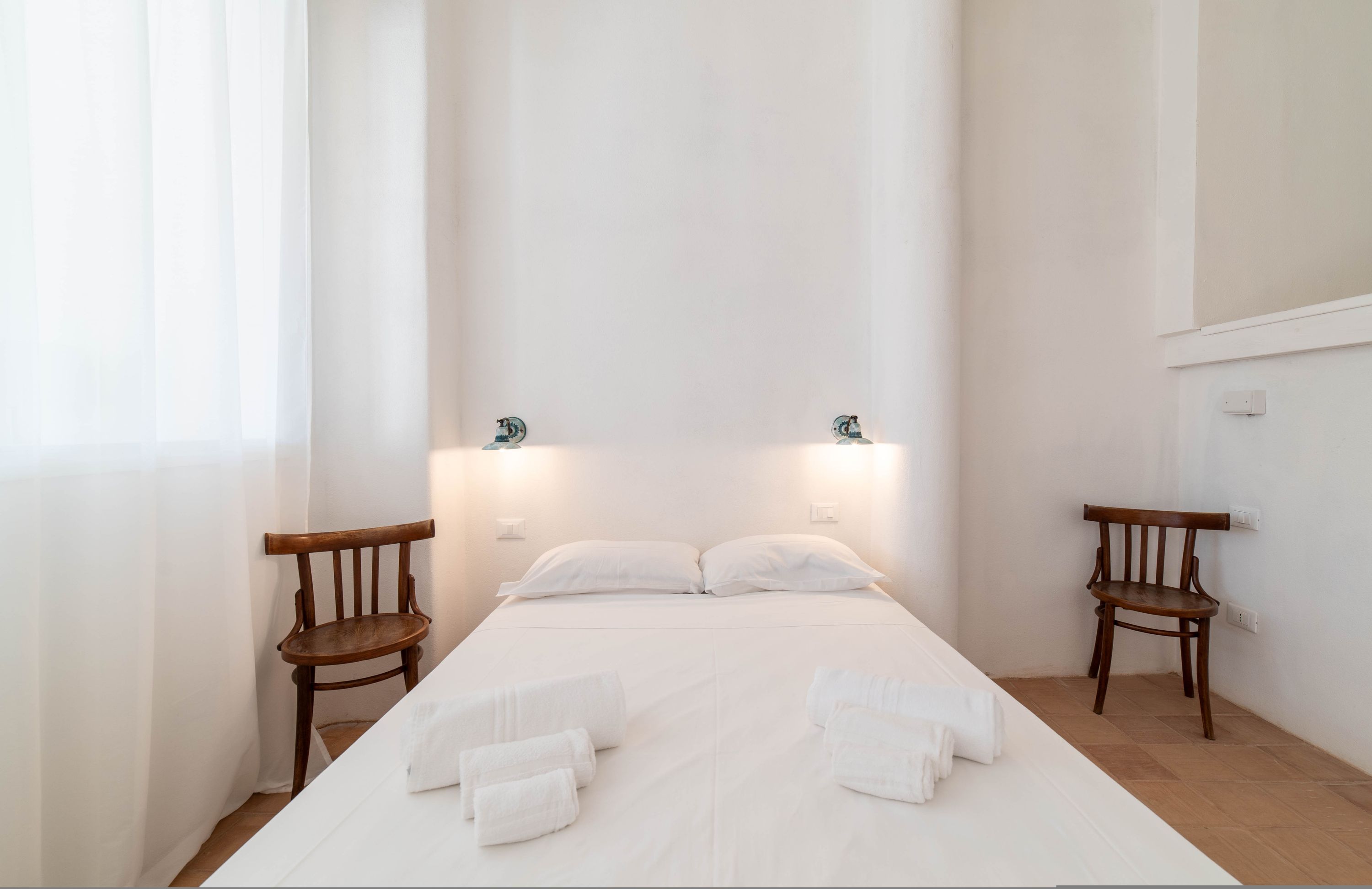 A minimalist hotel room with a large bed, two wooden chairs, and a bright, uncluttered space.
