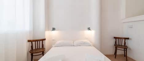 A minimalist hotel room with a large bed, two wooden chairs, and a bright, uncluttered space.