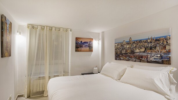 A hotel room with a large bed, a cityscape painting on the wall, and a window with sheer curtains. - Galea Studio by Wonderful Italy (Genoa)