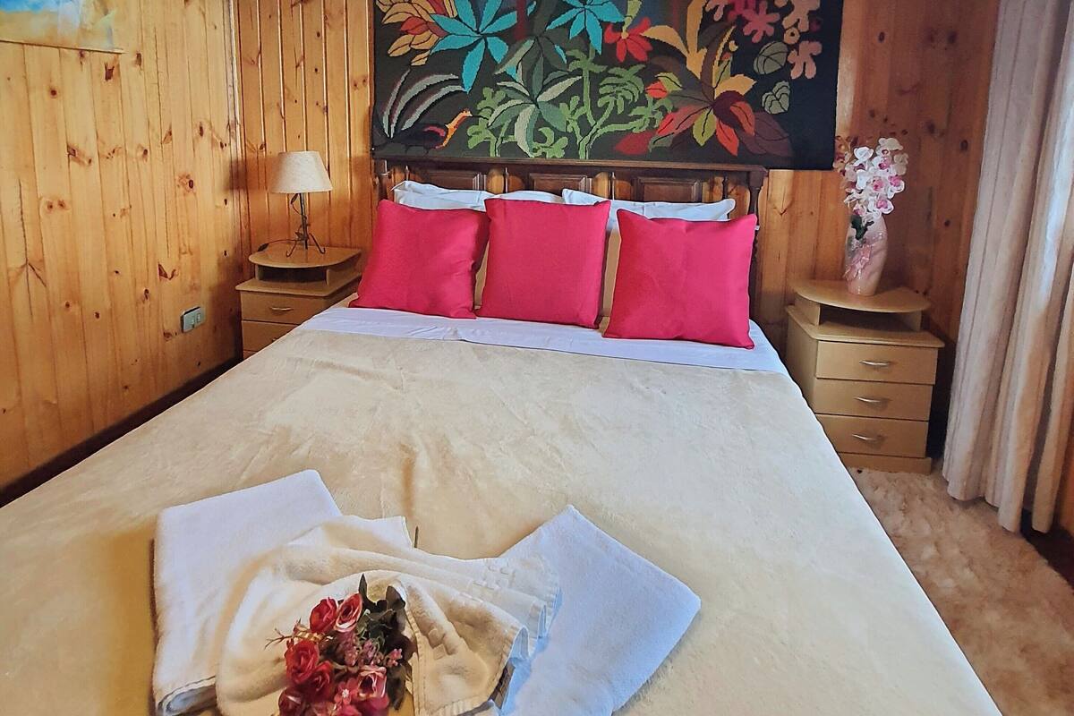 Una habitación de madera con una cama, almohadillas rojas, un tapiz floral, un jarrón con flores y una lámpara.