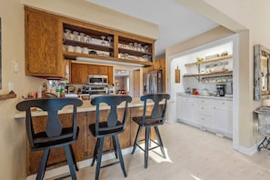 Une cuisine avec des meubles de rangement en bois, un four à micro-ondes et un réfrigérateur. On y trouve deux tabourets de bar et un comptoir sur lequel sont disposés divers objets.