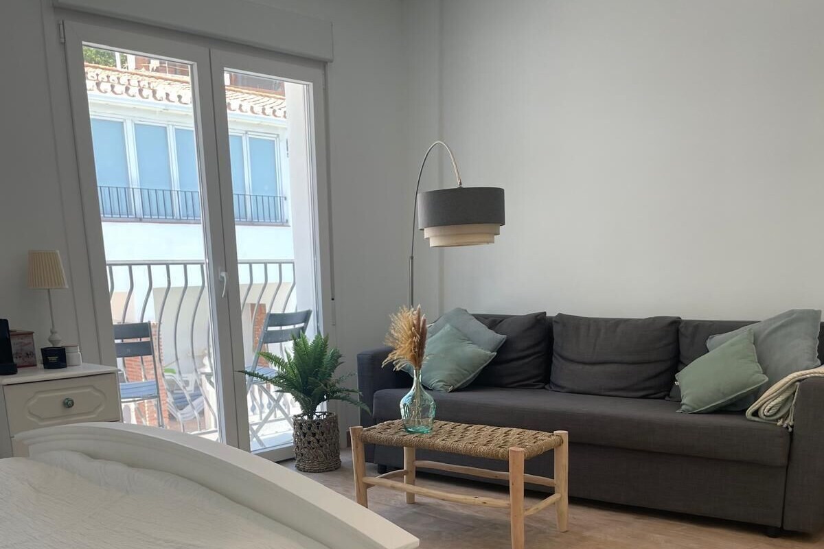 A modern bedroom with a bed, a grey sofa, a small wooden table, and a balcony with a railing.