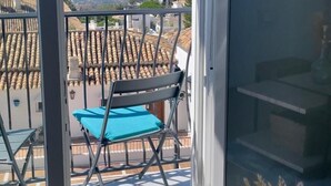 Un balcon donnant sur un paysage urbain, une chaise bleue et une table sur laquelle est posé un vase contenant des plantes séchées.
