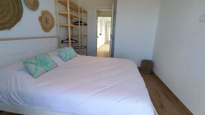 Une chambre à coucher avec un lit, des oreillers, une étagère en bois avec des cintres, un panier en osier et une porte donnant sur une autre pièce.