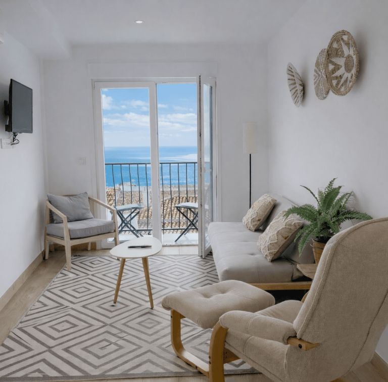 Un salon comprenant un canapé, un fauteuil et une petite table. Il y a un balcon avec vue sur la mer.
