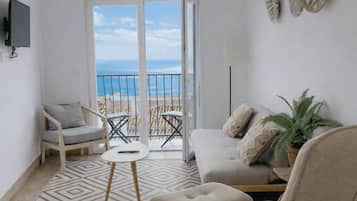 A living room with a sofa, armchair, and a small table. There is a balcony with a view of the sea.
