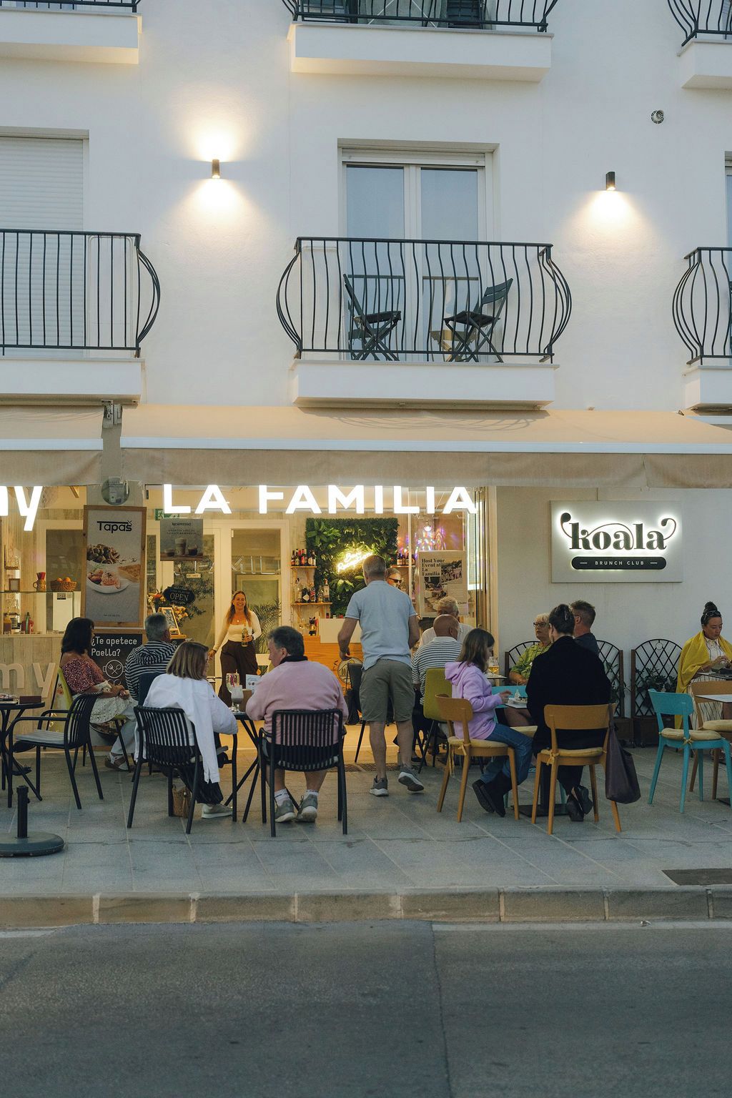 Un área de estar al aire libre de un restaurante con mesas y sillas, un letrero que dice 'La Familia' sobre la entrada y un letrero 'Koala' que indica un club de brunch.