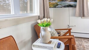 A room with a view of a sunset over a rocky landscape, a white vase with green plants, a book titled 'The Art of Home', and a remote control on a small table.