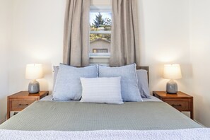 Una cama bien hecha con un cojín gris y blanco a rayas, dos lámparas grises sobre mesitas de noche de madera, y una ventana con cortinas.