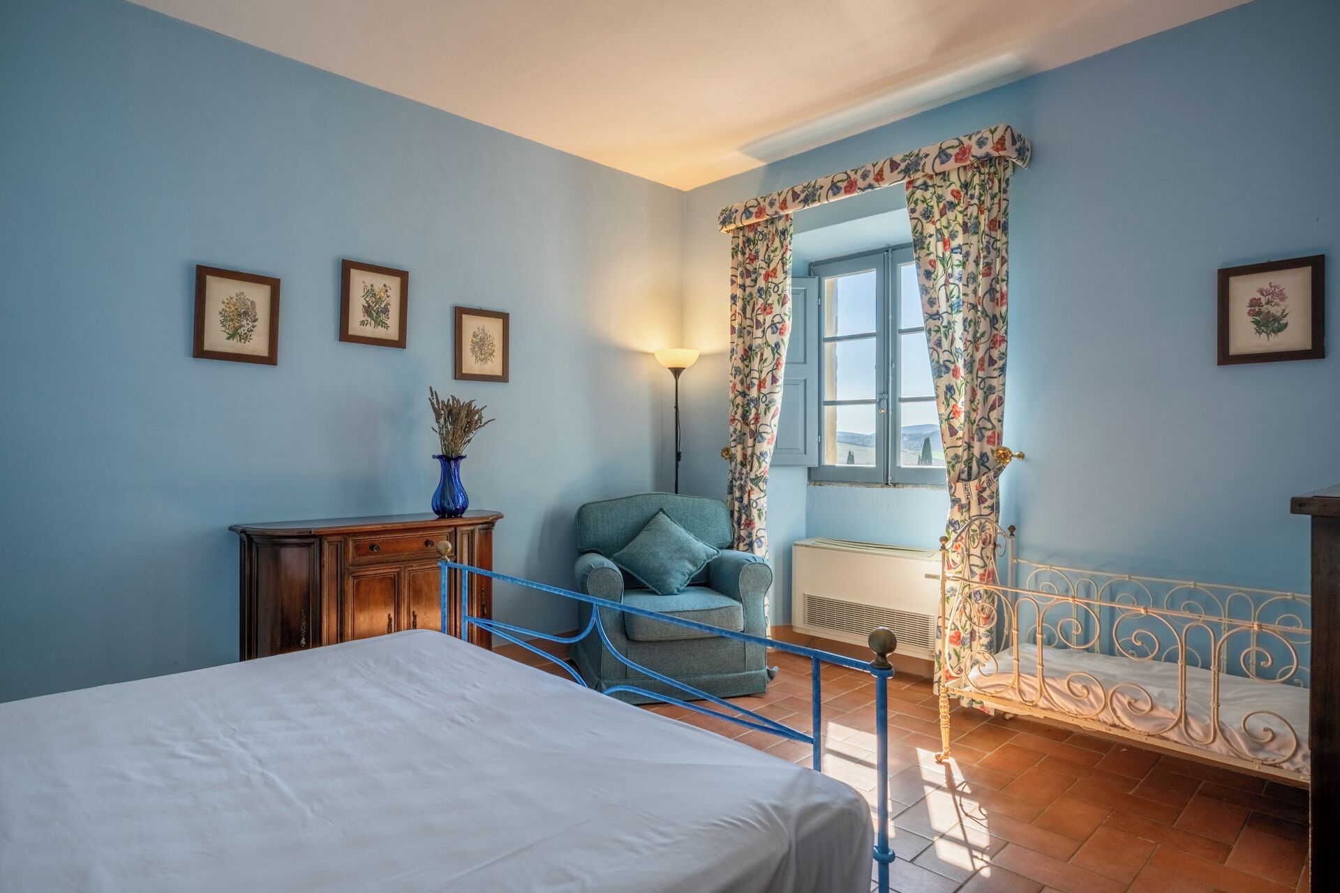 Een slaapkamer met een bed, een stoel, een dressoir en een raam met bloemen gordijnen.