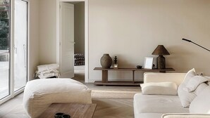 Ein modernes Wohnzimmer mit einer Couch, einem Sessel und einem Couchtisch. Eine Tür führt auf einen Balkon, und eine Lampe steht auf einem Beistelltisch.