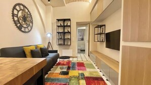 Un salon moderne avec une table basse en bois, un canapé noir agrémenté de coussins jaunes, un tapis coloré et une grande horloge murale.