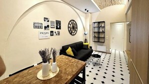 Un intérieur moderne avec un sol à carreaux noir et blanc, un canapé noir agrémenté de coussins jaunes, une table à manger en bois avec des vases, et un mur orné de tableaux encadrés et d’une horloge.