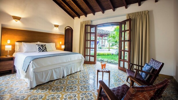 Habitación de hotel con cama, lámparas de noche, ventana con cortinas y vistas al jardín, además de una pequeña mesa con sillas.