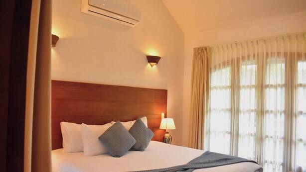 Une chambre d’hôtel avec une tête de lit en bois, un lit avec une literie blanche et grise, une lampe sur la table de chevet et une fenêtre avec des rideaux légers.