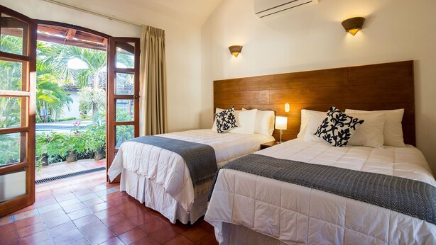 Une chambre d’hôtel avec deux lits, une tête de lit en bois, une fenêtre donnant sur un paysage verdoyant et un appareil de climatisation.