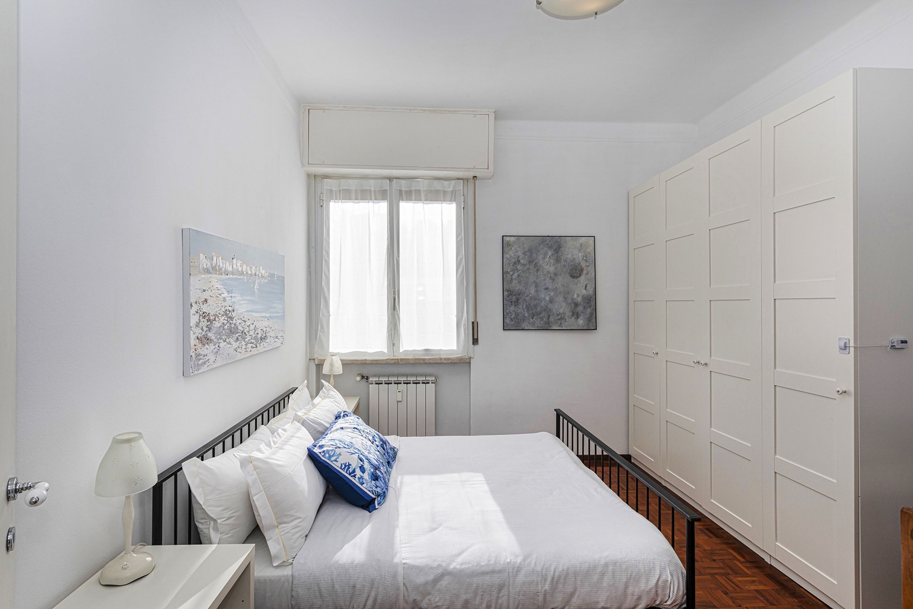 A bedroom with a bed, a nightstand, a window with curtains, a radiator, and a wardrobe.