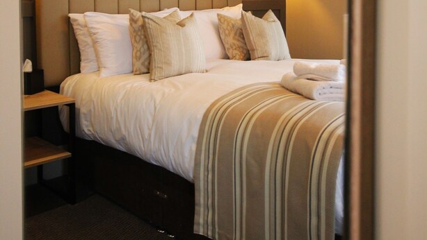 A hotel room with a large bed, headboard with wall sconces, a bedside table, and a carpeted floor. - The Coach & Horses (King's Lynn)
