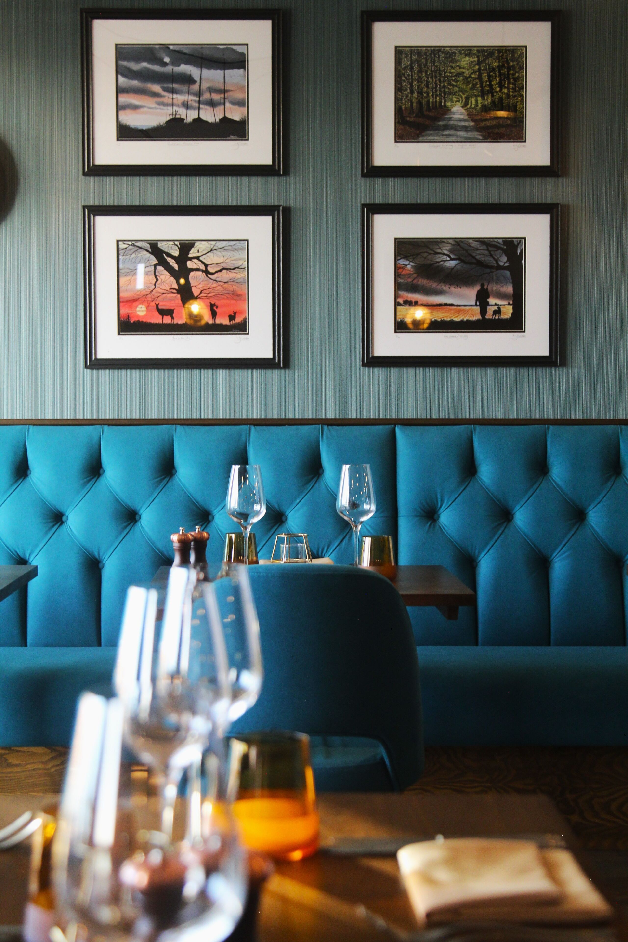 Un restaurante con un asiento tapizado en azul, obras de arte enmarcadas en la pared y una mesa servida con copas de vino y una botella.