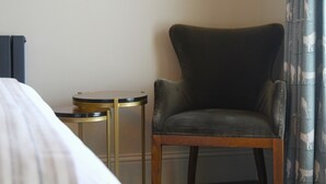 A hotel room with a bed, a dark leather armchair, a small round table with a gold frame, and a curtain with a pattern. - The Coach & Horses (King's Lynn)