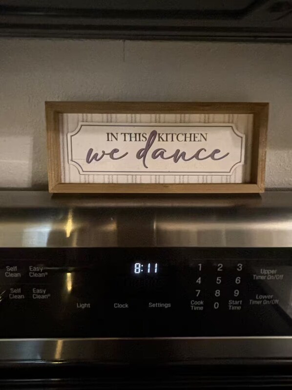 A kitchen with a digital clock displaying 8:11 and a sign above it that reads 'IN THIS KITCHEN we dance.'
