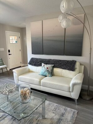 A modern living room with a white leather sofa, a glass coffee table, a large abstract painting, and a floor lamp.