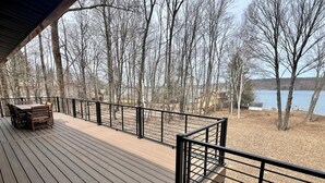 Eine Holzterrasse mit Tisch und Stühlen, mit Blick auf einen See, Bäume und ein Gebäude im Hintergrund.