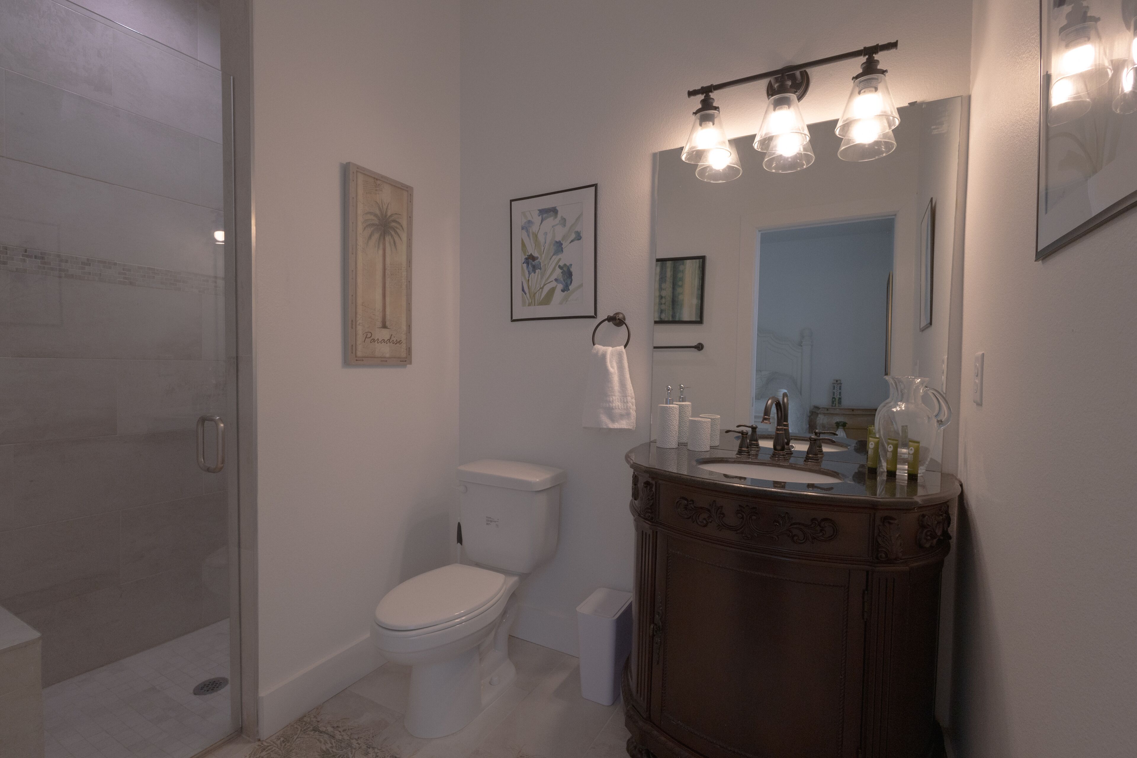 Un baño con cabina de ducha de vidrio, inodoro blanco, tocador de madera con espejo e iluminación en la pared.