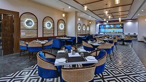 A spacious dining area with round tables and blue upholstered chairs, featuring a patterned floor and a large screen in the background.