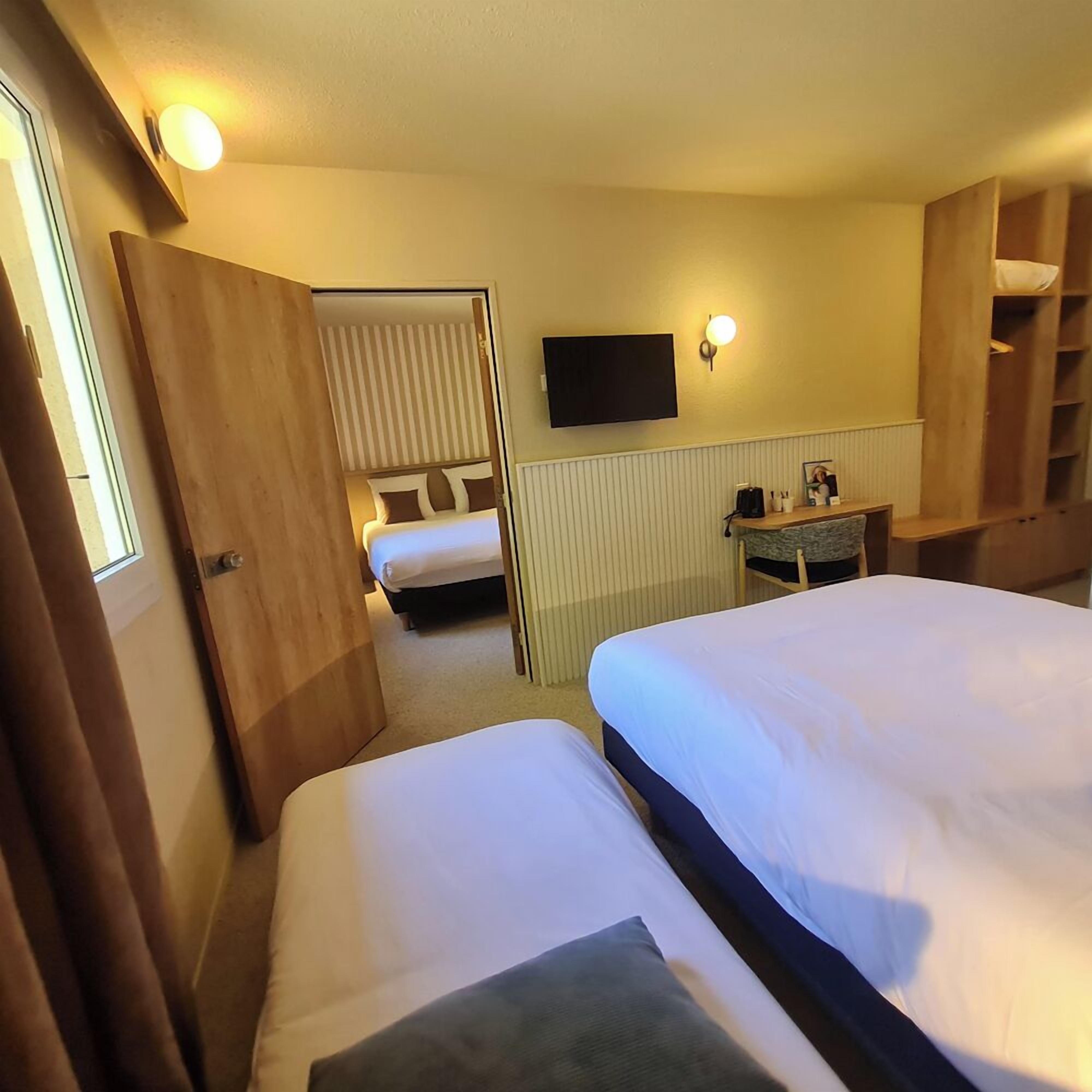 Camera d'albergo con due letti, una TV a muro, un tavolino con una sedia e una scaffalatura in legno.