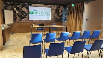 Een vergaderruimte met een groot scherm waarop 'BRIT HOTEL ALENÇON' staat, en een rij blauwe stoelen die naar het scherm gericht zijn.