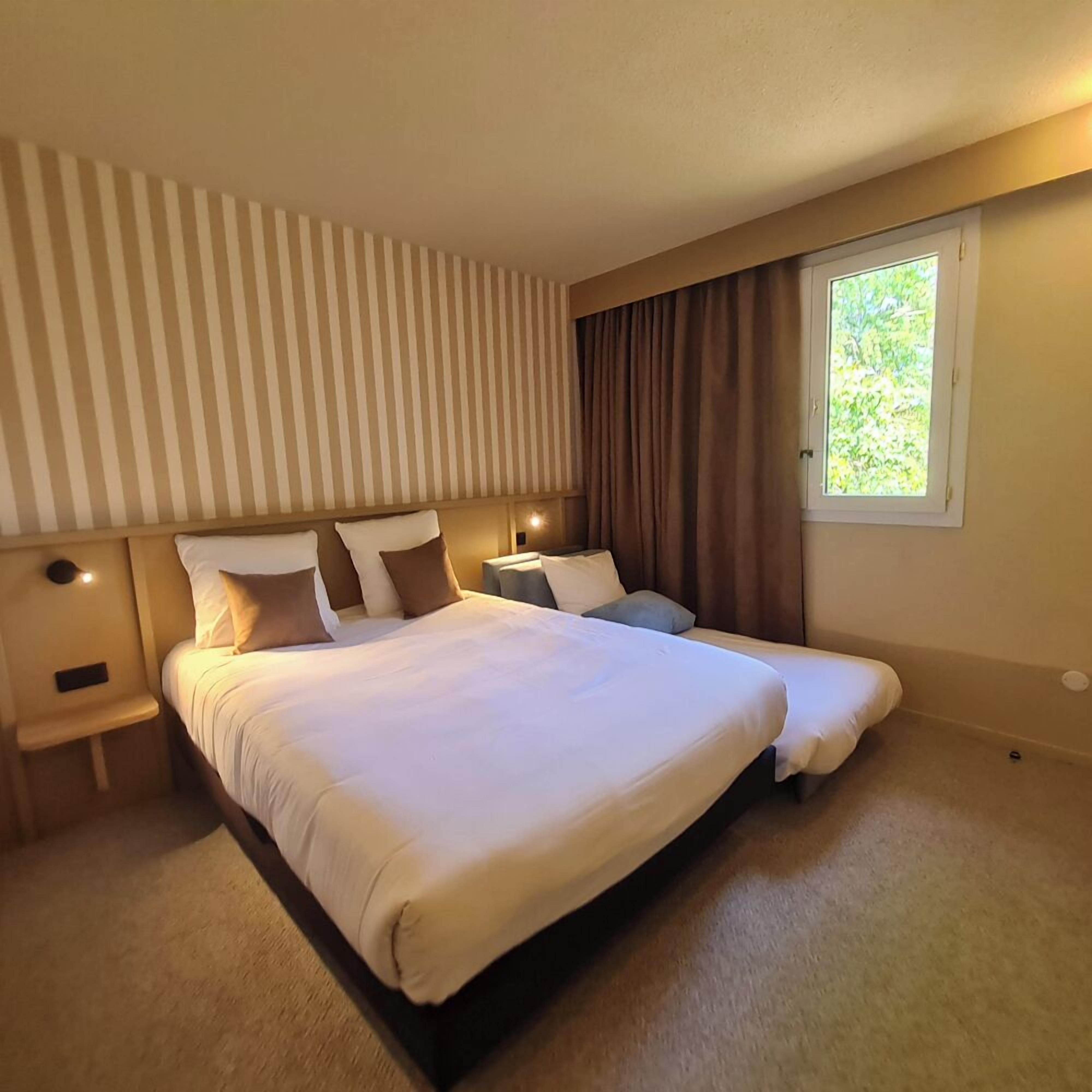 A hotel room with a large bed, a bedside table, a nightstand, a window with curtains, and a striped wall.