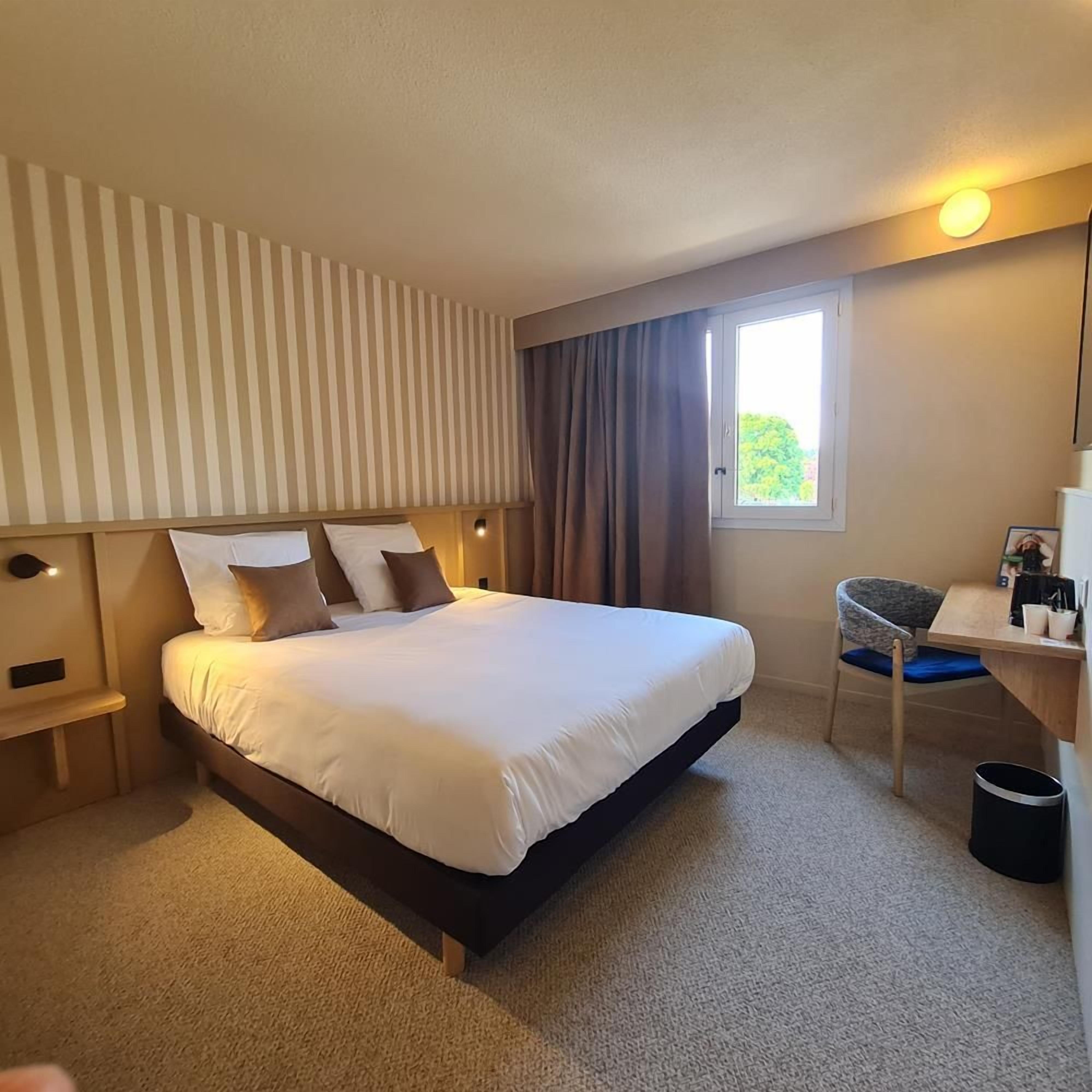 Une chambre d’hôtel avec un grand lit, un bureau, une chaise, une fenêtre avec des rideaux et un luminaire fixé au mur.