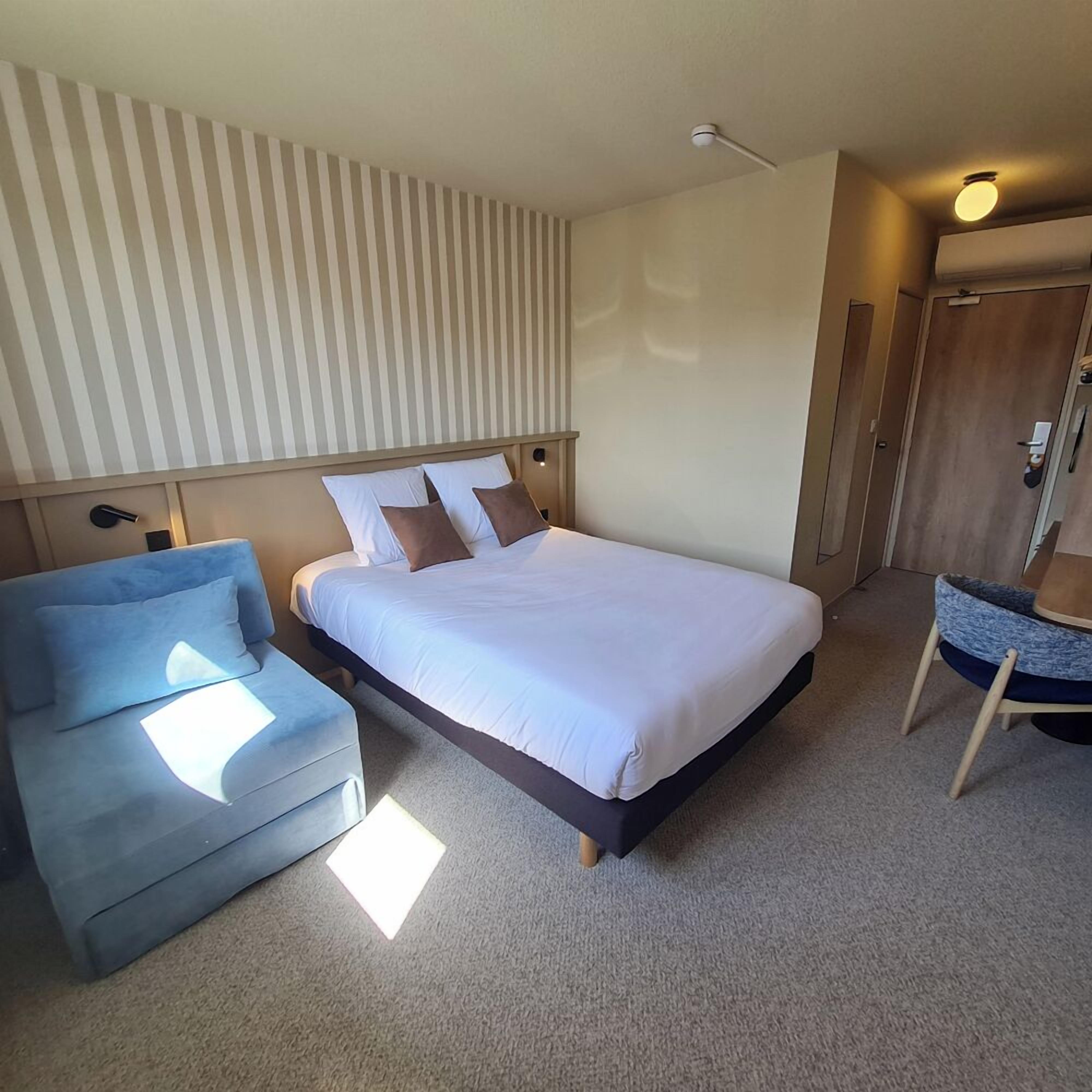 Un dormitorio con una cama, un sillón azul y una pequeña zona de estar con una silla y una mesa redonda.