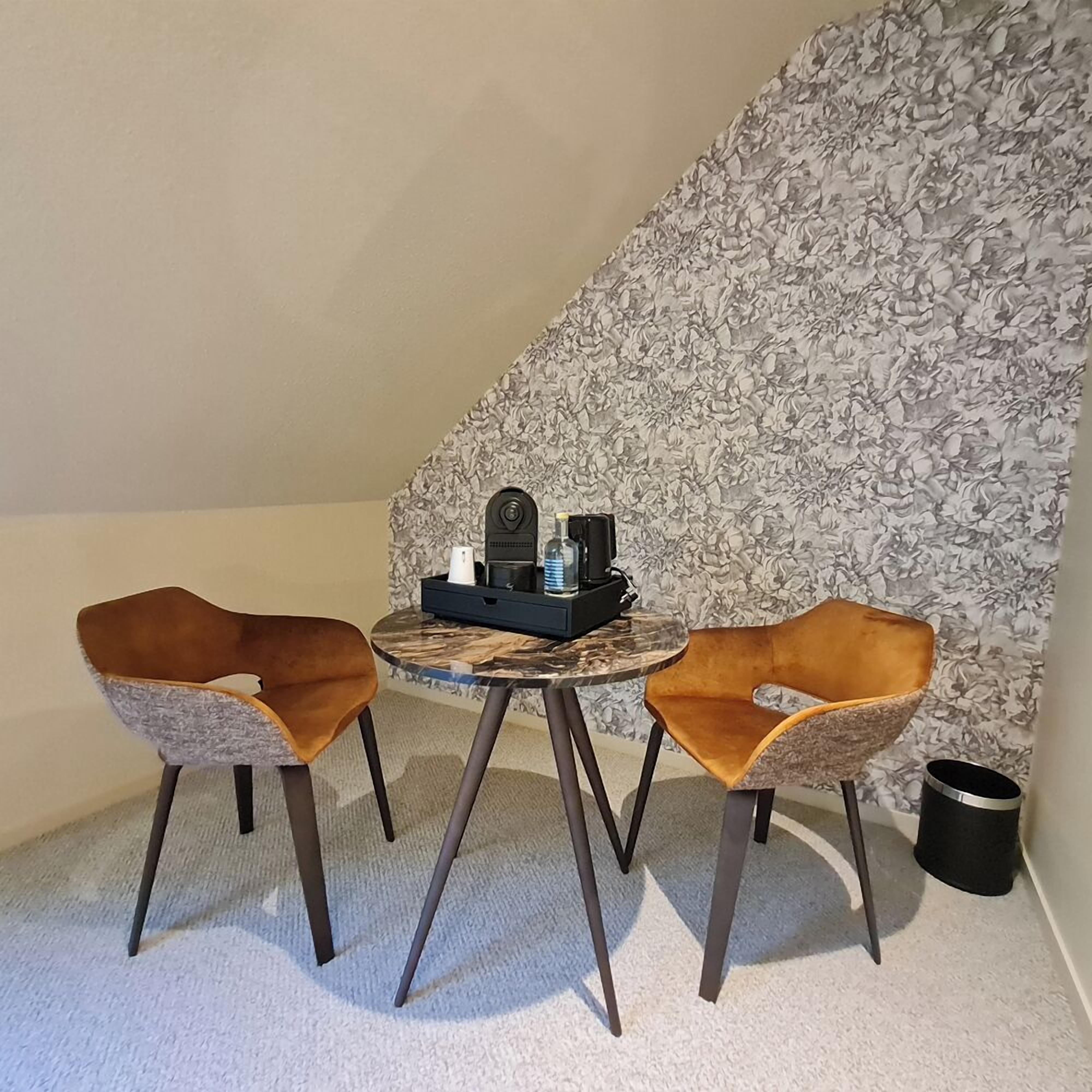 Ein kleiner Raum mit einem runden Tisch, zwei Stühlen, einer schwarzen Mülltonne und einer Wand mit floralem Muster.