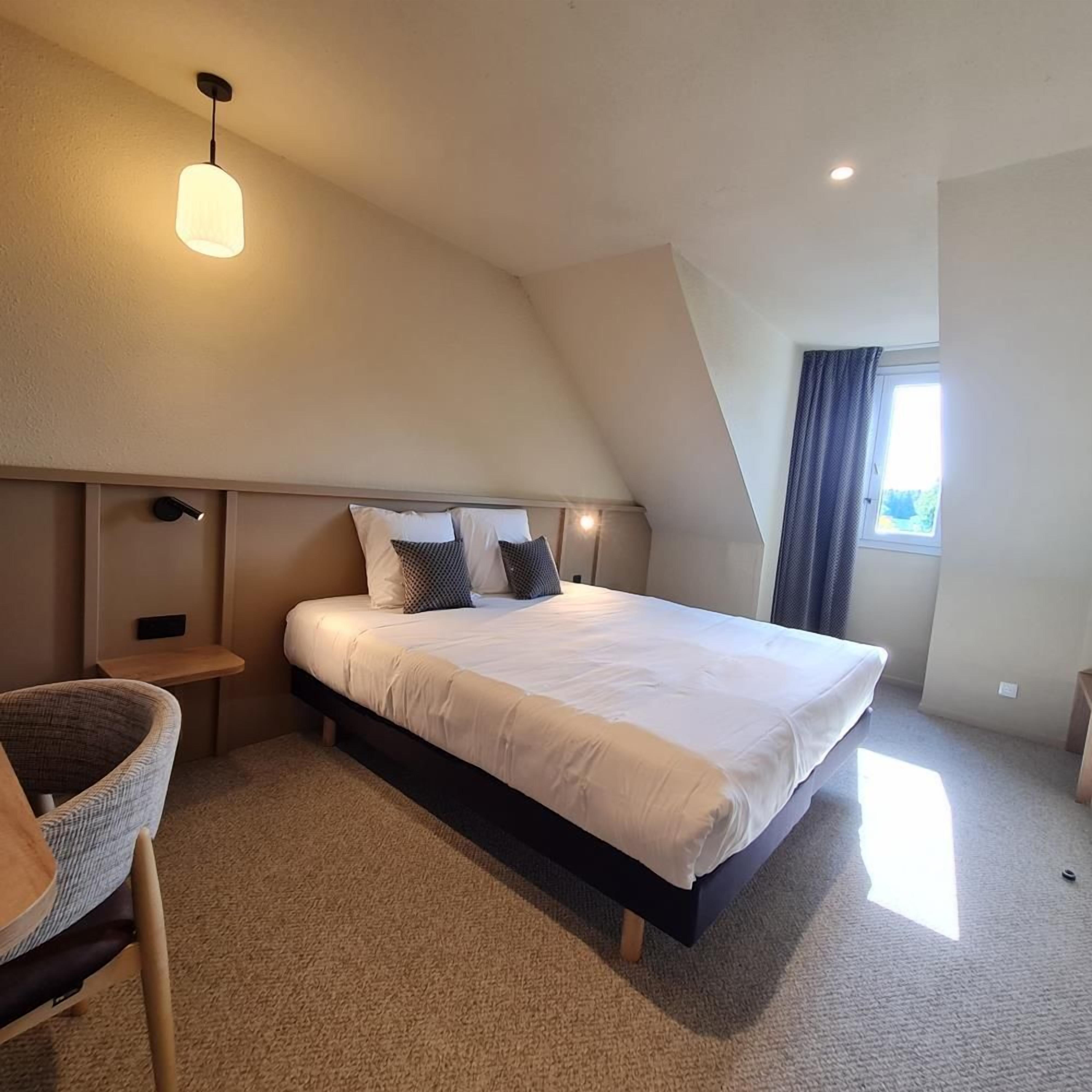 A bedroom with a large bed, a bedside table, a chair, and a window with curtains.