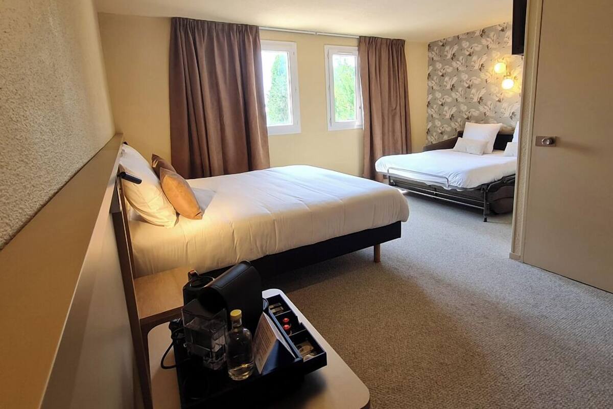 A hotel room with two beds, a desk with a coffee maker, and a small table with a bottle and glasses.
