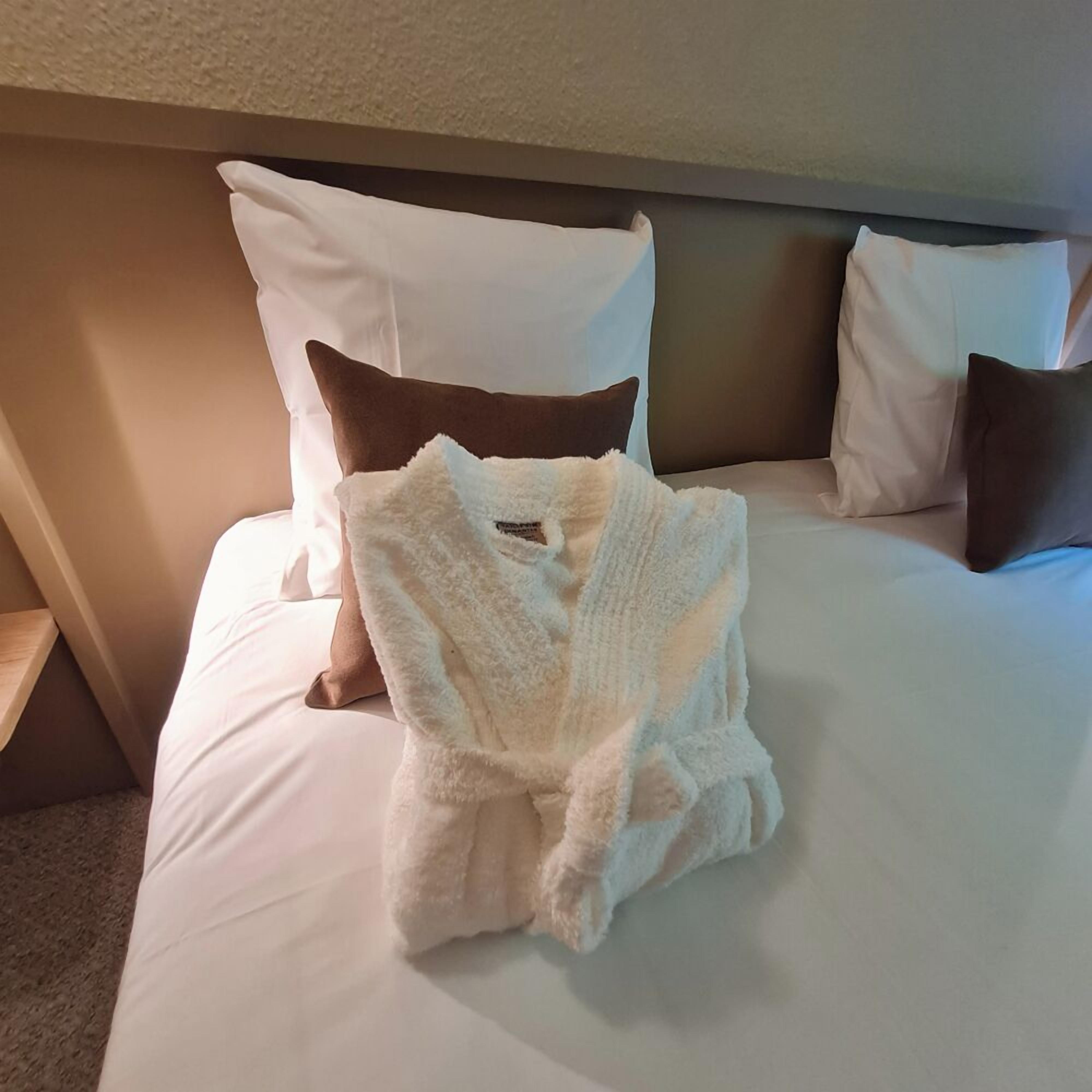 A neatly made bed with white linens, a fluffy white bathrobe, and two brown pillows.