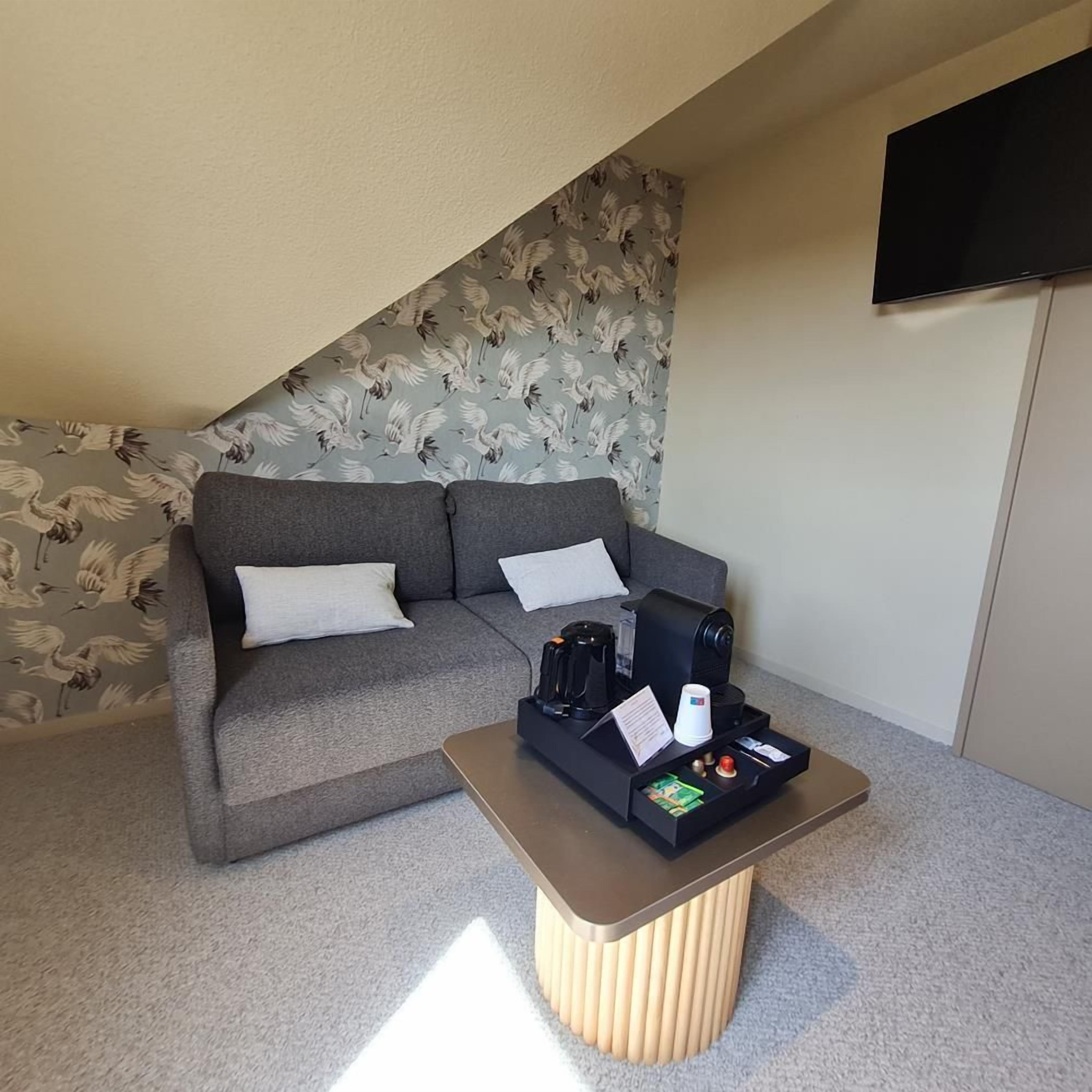 A room with a grey sofa, a coffee table with a kettle, and a flat-screen TV mounted on the wall.