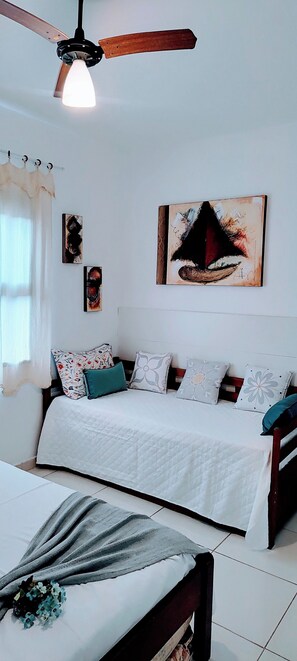 A bedroom with a bed, pillows, a bedside table, a ceiling fan, and artwork on the wall. - Guarujá Sol e Praia (Guarujá)